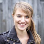 Portrait of a young woman smiling in a leather jacket against a wooden fence background outdoors.